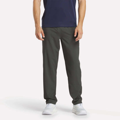 A man stands facing forward against a plain white background wearing Reebok Men's Train Woven Unlined Pants, a navy shirt, and white sneakers. The image shows him from shoulders to feet—ideal attire for gym workouts.