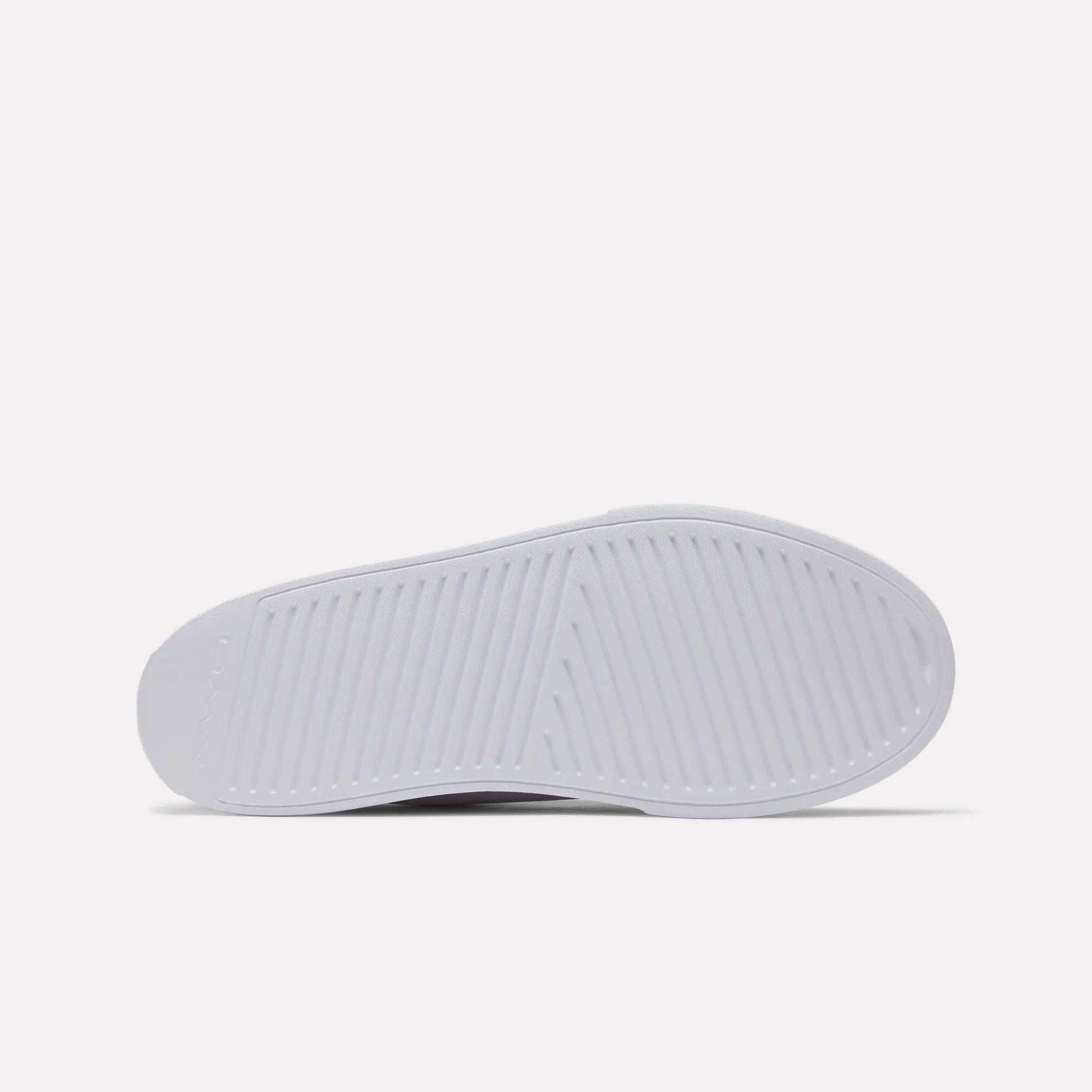 The image shows the sole of a white Women's OnLux Court Lite Walking Shoe by Reebok, viewed from the bottom. The sole has a textured pattern with diagonal and crosswise grooves for traction. The background is plain and light-colored.