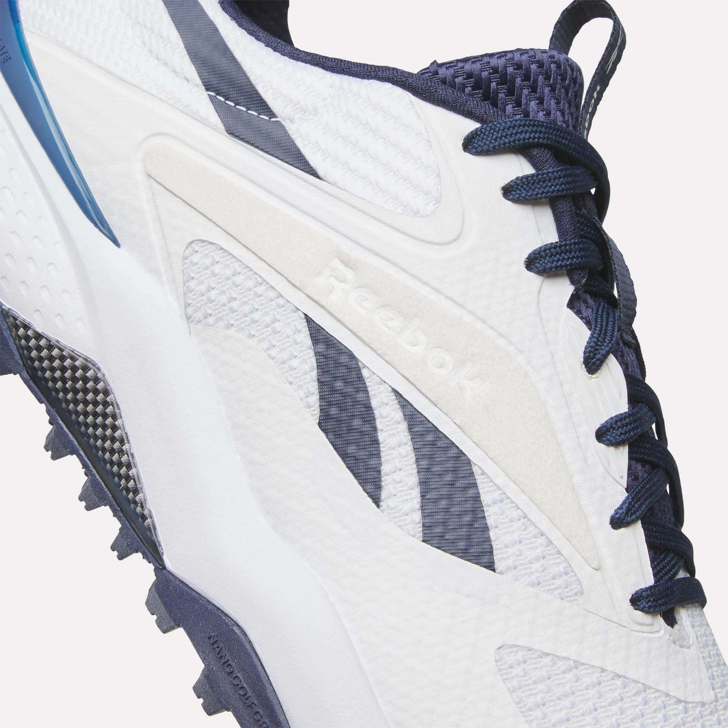 Close-up of Reebok Nano Golf Shoes in white and navy, featuring a lightweight FlexWeave upper, blue and black accents, dark blue laces, and visible Reebok branding on the side.