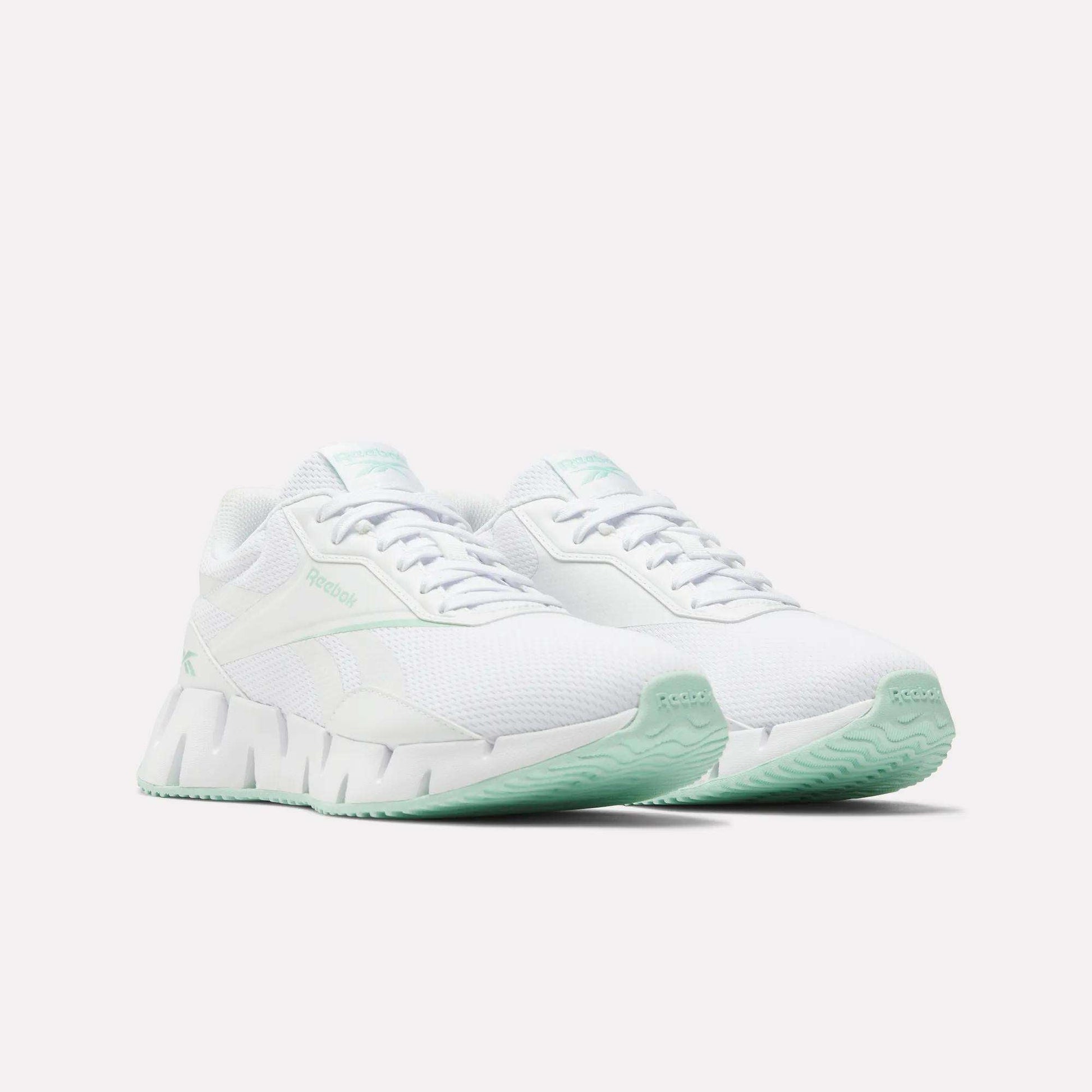 A pair of Reebok Women's Zig Dynamica STR Shoes in white, featuring breathable mesh and light mint green accents on the soles and branding, displayed on a white background.