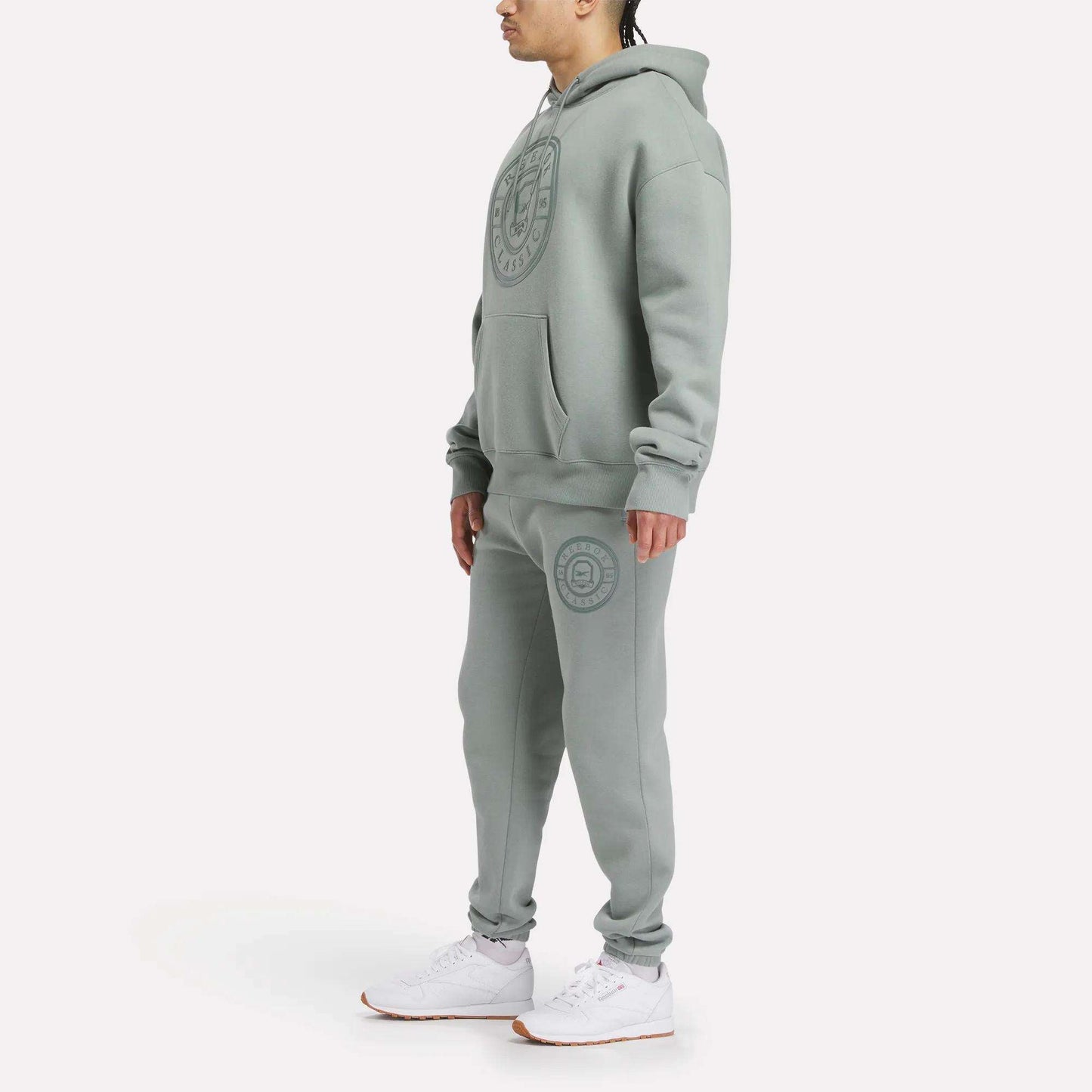 A person stands wearing a relaxed fit Reebok Men's Crest Hoodie and sweatpants set in light gray fleece with large circular logos, paired with white sneakers, against a plain white background.