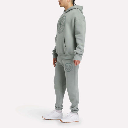 A person stands wearing a relaxed fit Reebok Men's Crest Hoodie and sweatpants set in light gray fleece with large circular logos, paired with white sneakers, against a plain white background.
