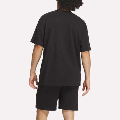 A person with curly hair is shown from the back against a plain white background, wearing a loose black Reebok Men's Crest T-Shirt and black shorts for everyday comfort.