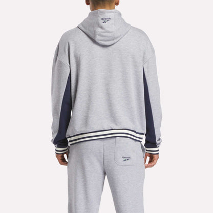A person wearing a light gray Reebok Men's Lawn Club French Terry Hoodie, seen from behind. This hoodie features dark blue sleeve accents and striped cuffs and waistband.