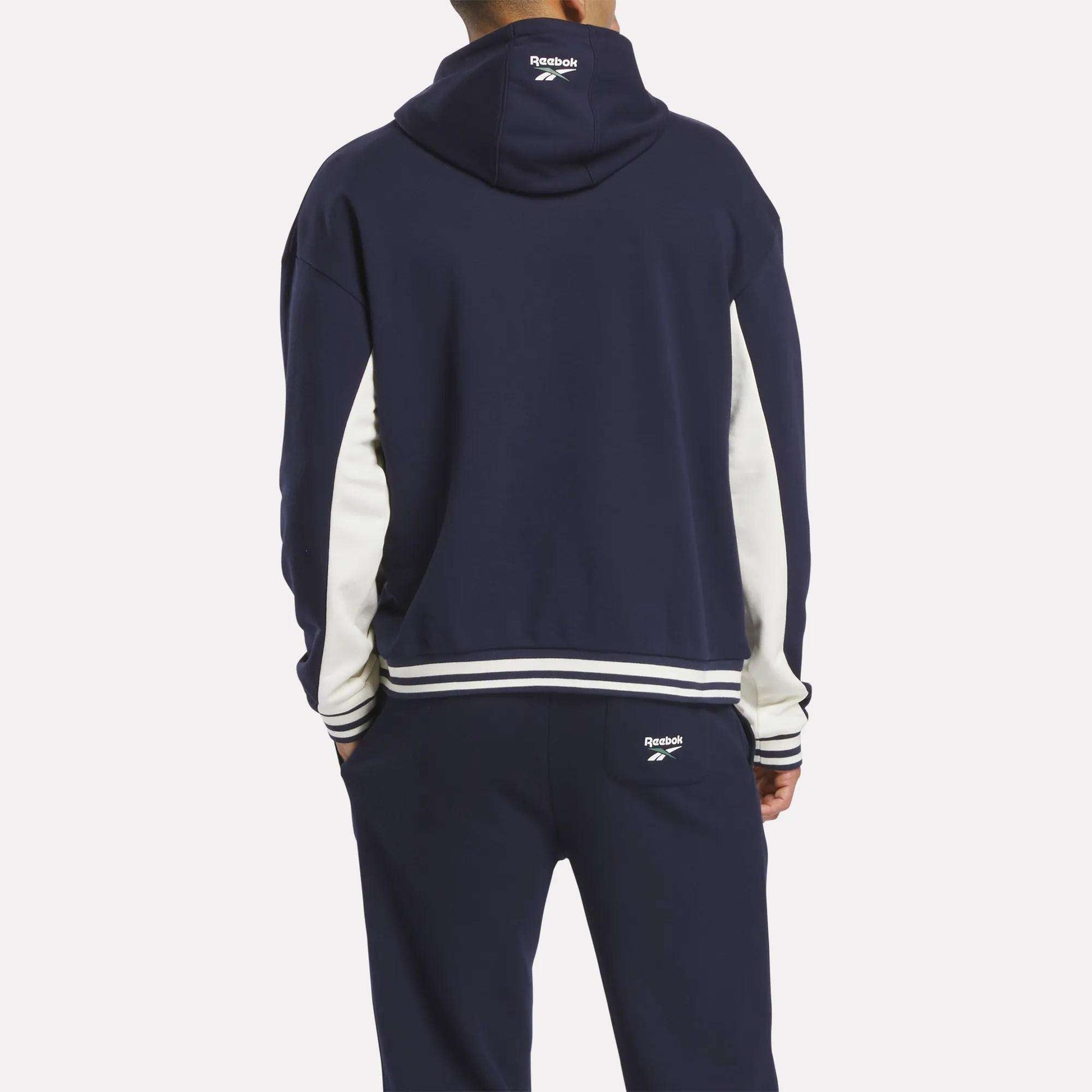 A person faces away wearing the Reebok Men's Lawn Club French Terry Hoodie tracksuit in navy blue with white sleeve stripes and trim. The cotton-blend hoodie displays the Reebok logo on the hood's back and pants pocket.