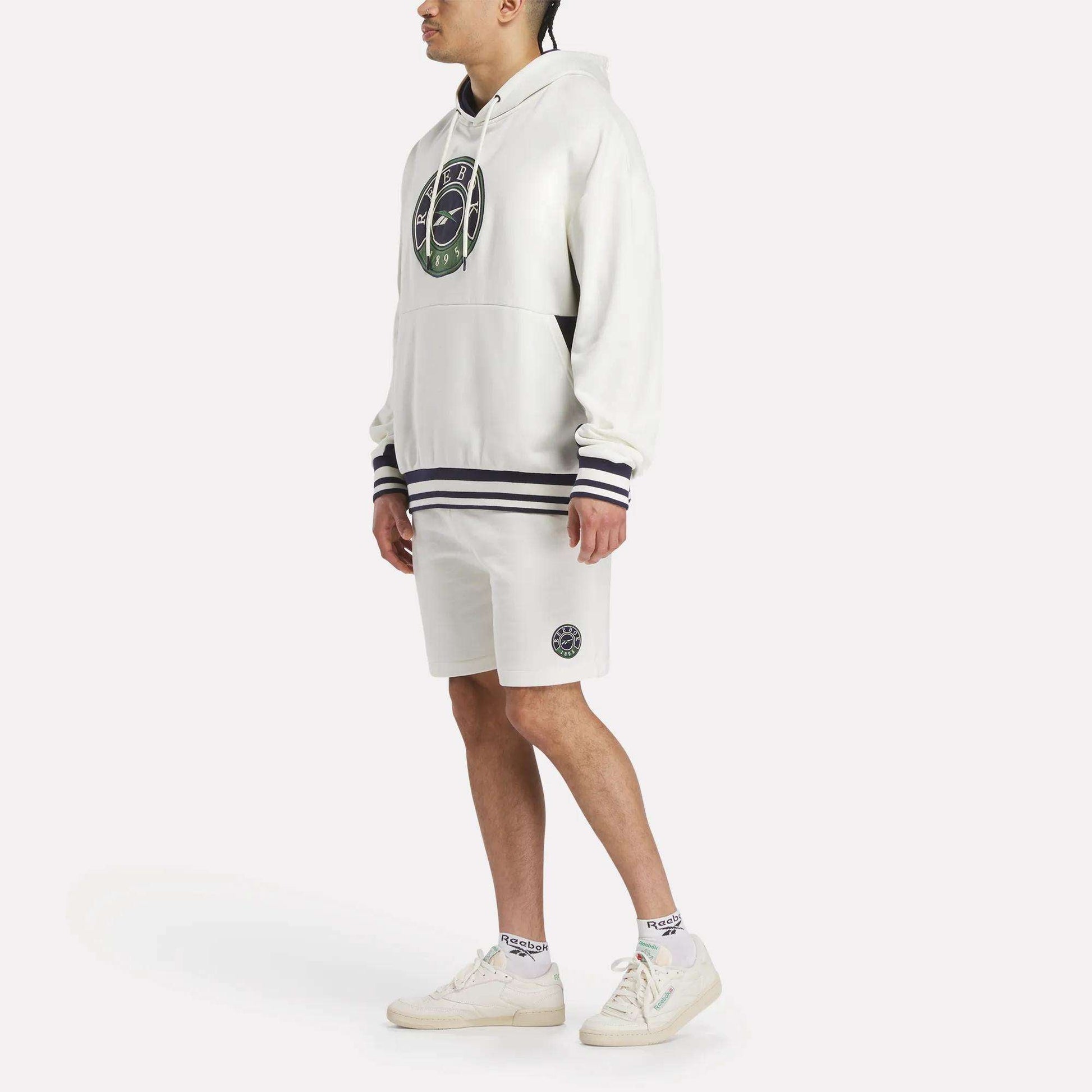 A man wears the Reebok Men's Lawn Club French Terry Hoodie featuring a circular green and black logo, paired with matching white shorts, white socks, and sneakers, all set against a plain background.