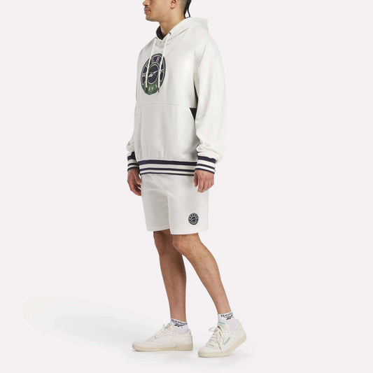 A man wears the Reebok Men's Lawn Club French Terry Hoodie featuring a circular green and black logo, paired with matching white shorts, white socks, and sneakers, all set against a plain background.