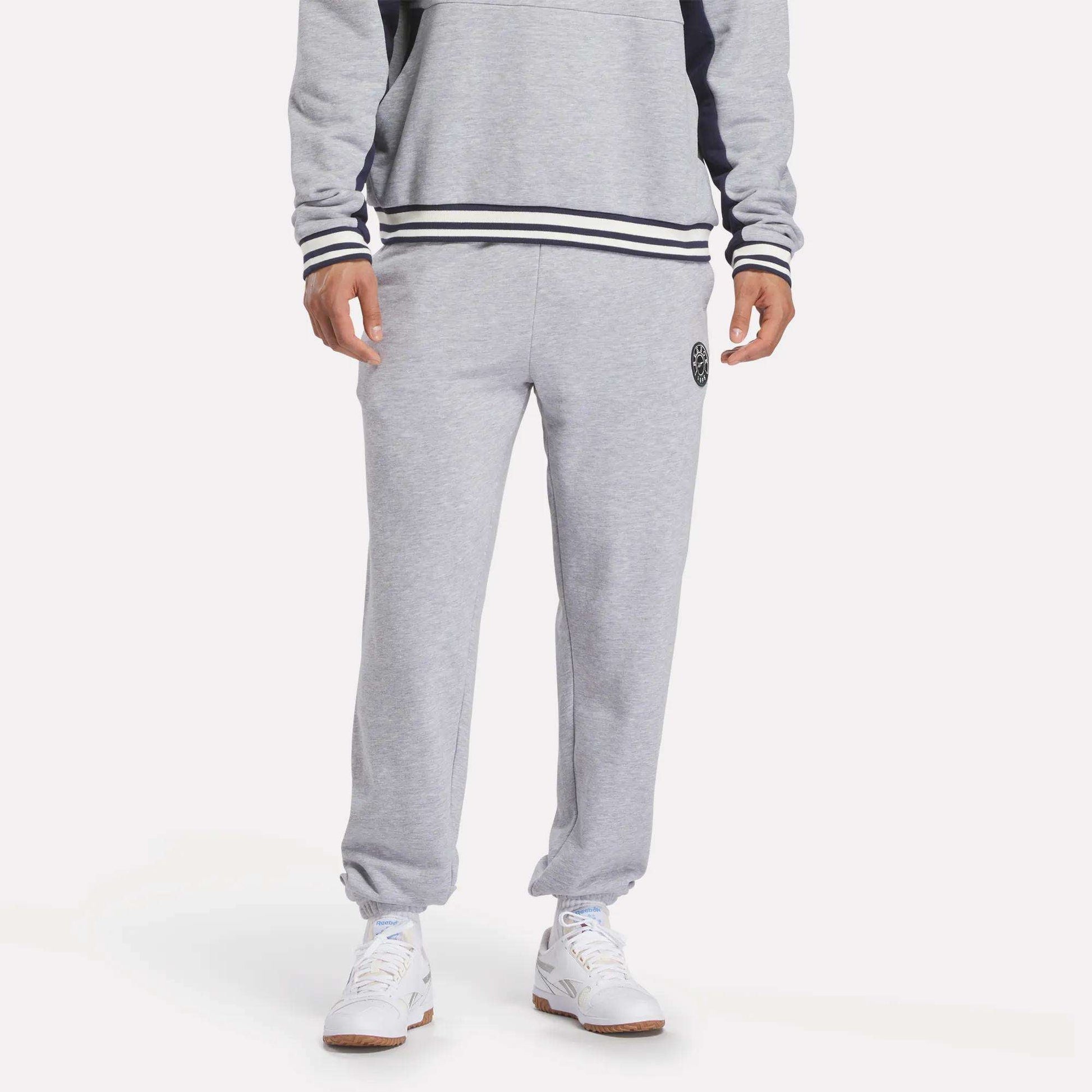A person in Reebok Men's Lawn Club French Terry Jogger Pants and a matching gray sweatshirt with black and white striped cuffs and hem, paired with white sneakers, stands against a plain white background.