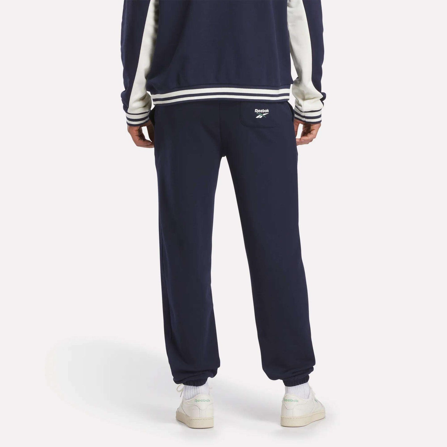 A person stands facing away, wearing Reebok Men's Lawn Club French Terry Jogger Pants in navy with a white logo on the back pocket, white sneakers, and a matching navy jacket—classic sporty style crafted from a comfortable cotton-blend.