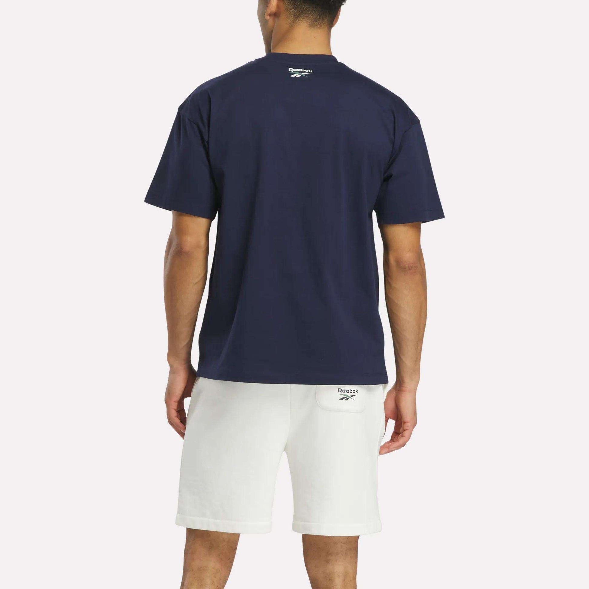A person faces away, wearing a navy blue Reebok Men's Lawn Club T-Shirt made of luxe cotton and white shorts. The Lawn Club logo is on the back of the shirt near the neck and on the waistband of the shorts.