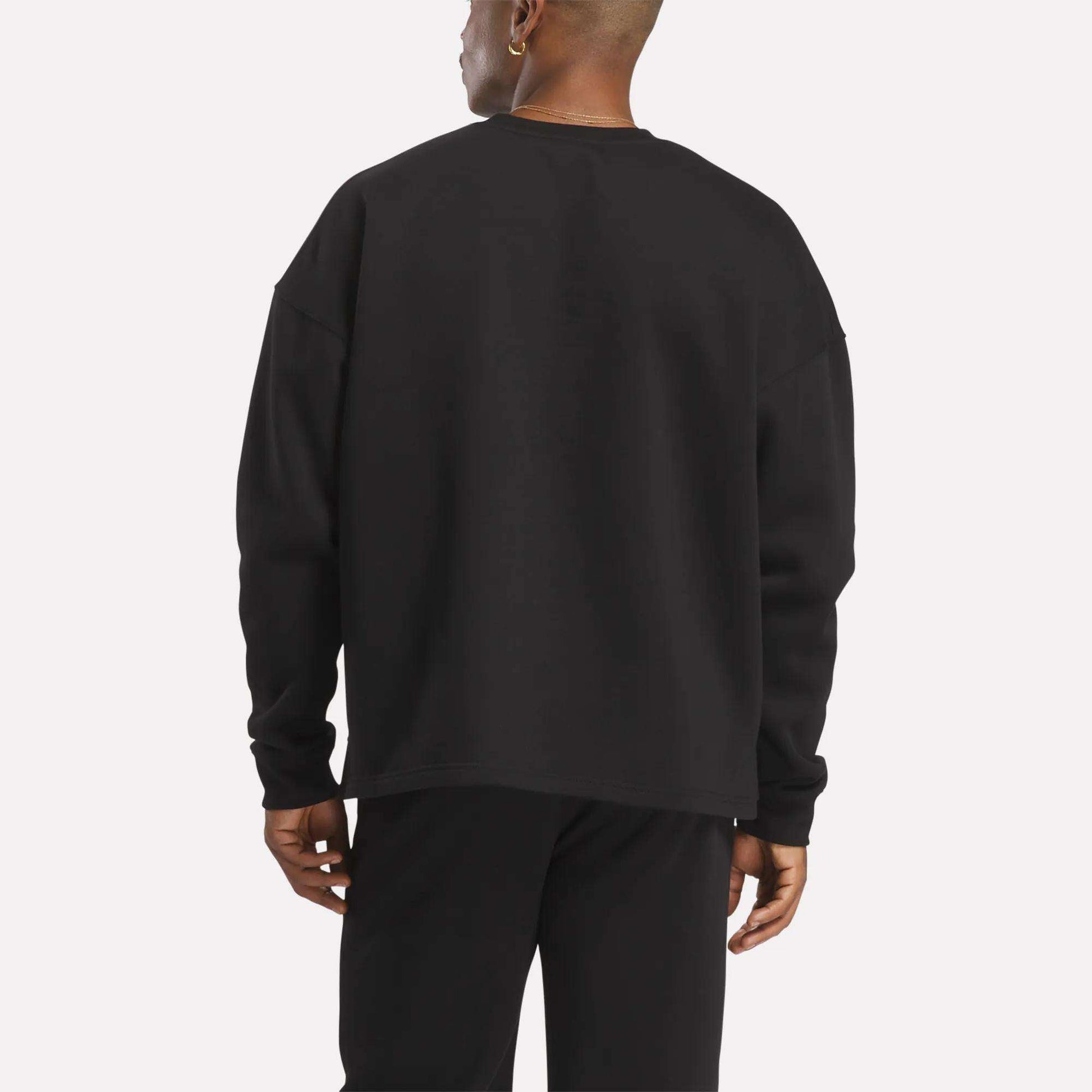 Facing away from the camera against a plain, light background, a person wears Reebok's Men's Wardrobe Essentials Sweatshirt with black pants, featuring a relaxed fit and crafted from soft cotton-blend fleece.