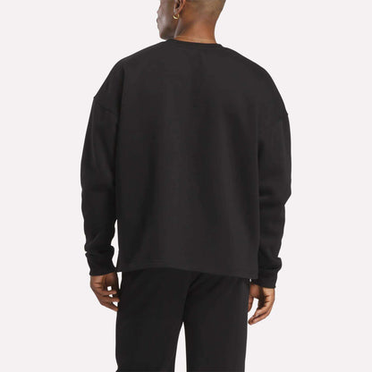 Facing away from the camera against a plain, light background, a person wears Reebok's Men's Wardrobe Essentials Sweatshirt with black pants, featuring a relaxed fit and crafted from soft cotton-blend fleece.