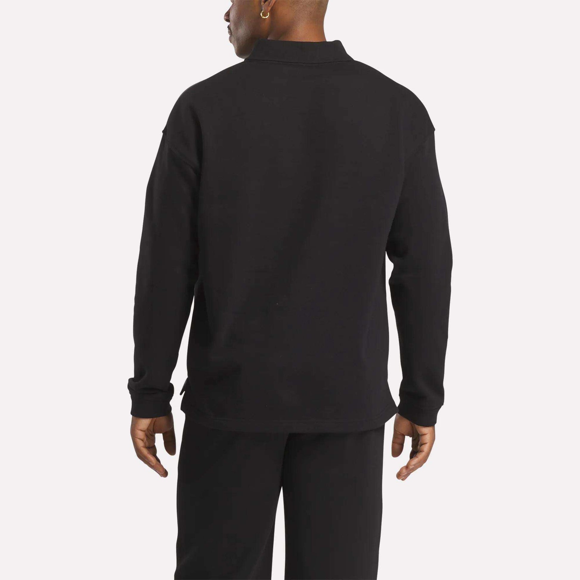 A person faces away from the camera in a Reebok Men's Wardrobe Essentials Long Sleeve Polo Shirt and black pants, highlighting the relaxed fit of this cotton-blend polo against a plain white background.