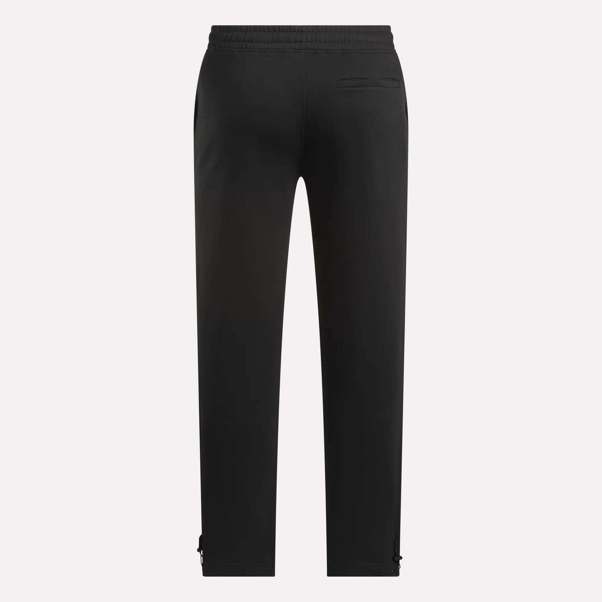 The Reebok Men's Wardrobe Essentials Pants are black, shown from the back with an elastic waistband, right-side back pocket, straight legs with small adjustable ankle straps, and made from soft cotton-blend fleece. Background is plain and light.