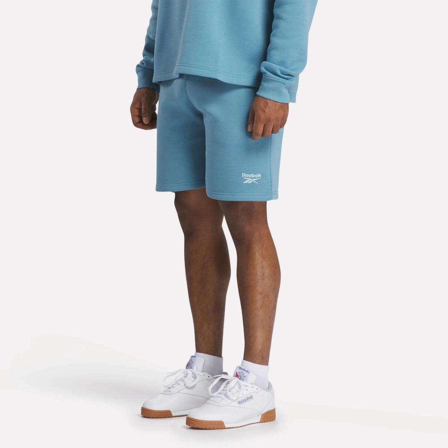 A person stands against a plain, light background wearing light blue relaxed fit sweatshirt, Reebok Men's Wardrobe Essentials Shorts, and white Reebok sneakers with gum soles, showing only the lower half of their body.