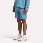 A person stands against a plain, light background wearing light blue relaxed fit sweatshirt, Reebok Men's Wardrobe Essentials Shorts, and white Reebok sneakers with gum soles, showing only the lower half of their body.