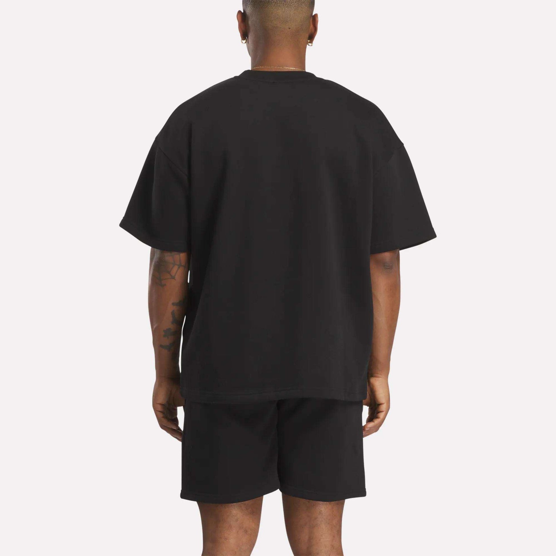 A person stands with their back to the camera, wearing a relaxed fit Men's Wardrobe Essentials T-Shirt by Reebok in black. The plain light background emphasizes the outfit, and their arms hang naturally at their sides.