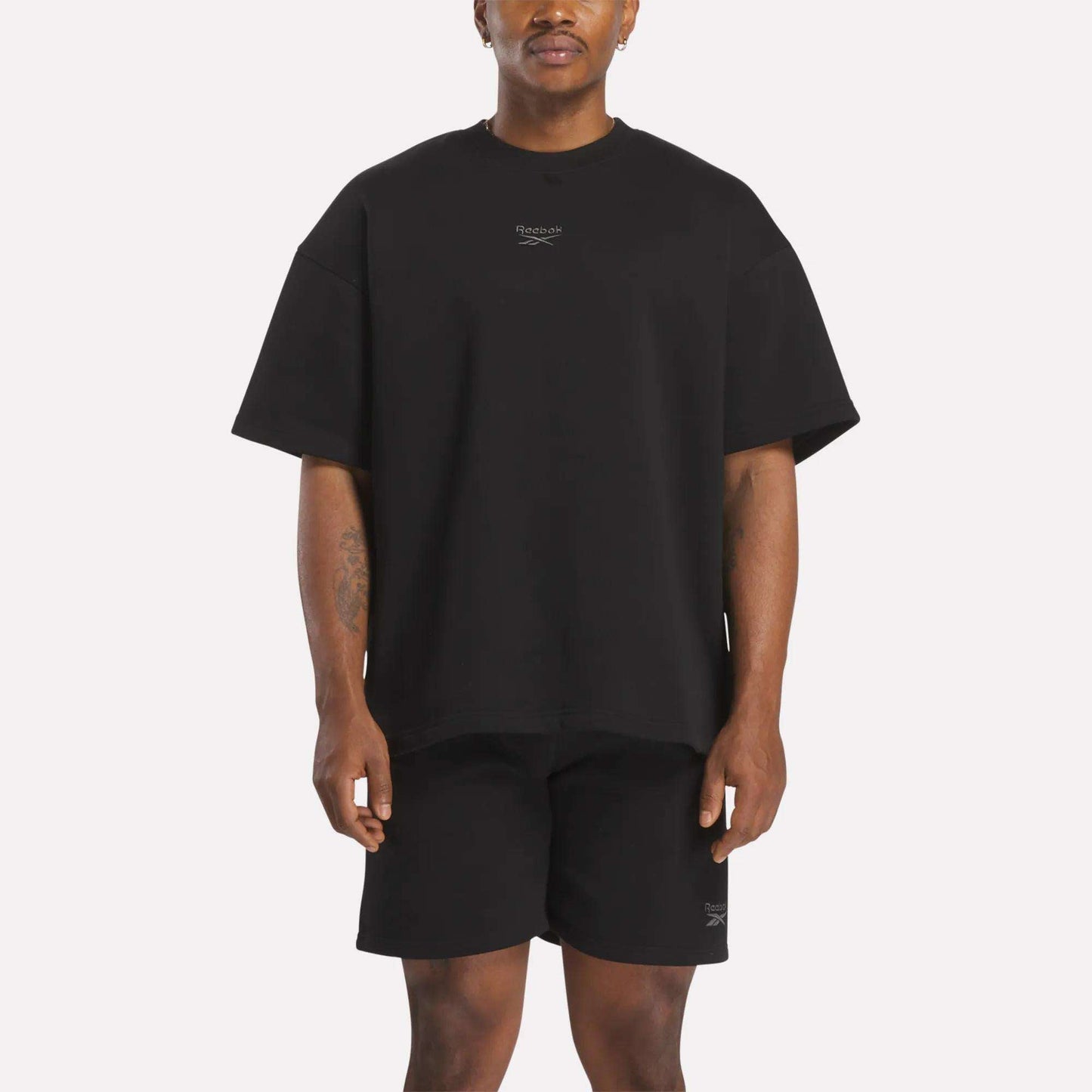 A man faces forward in a relaxed fit black Reebok Men's Wardrobe Essentials T-Shirt and black shorts, both featuring small white logos on the chest and left leg. The background is plain white.
