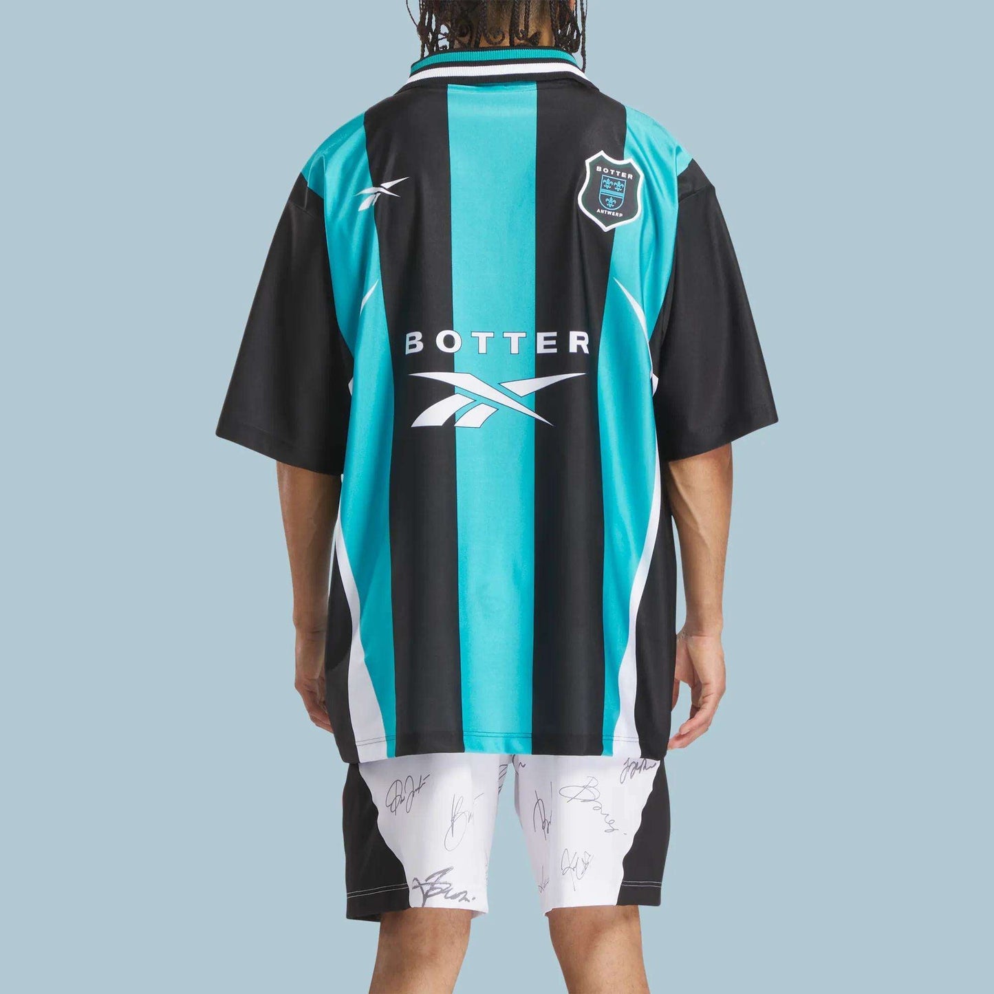 A person stands facing away, wearing the Reebok x Botter LTD Reverse Soccer Men's Tee—black and teal vertical stripes with white accents and logos by Reebok—and white shorts with autographs, set against a light blue background.