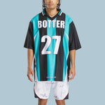 A person wears the Reebok x Botter LTD Reverse Soccer Men's Tee in teal and black stripes with BOTTER and 27, paired with signed white shorts. This standout Reebok look pops against a light blue backdrop.