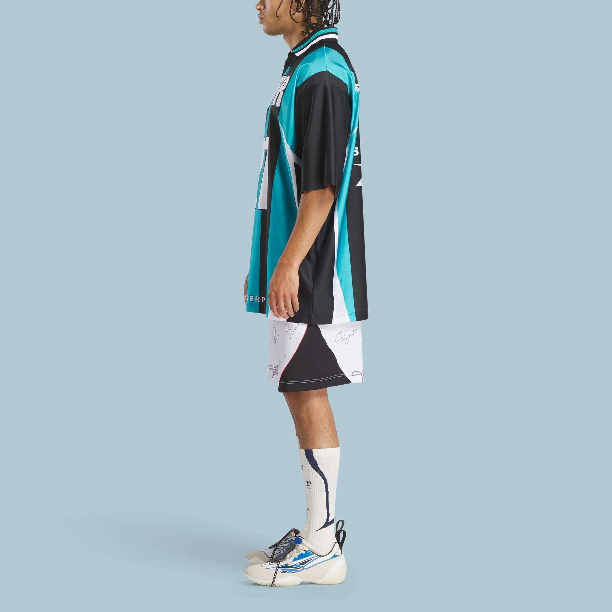 A person stands in profile against a light blue background, wearing the Reebok x Botter LTD Reverse Soccer Men's Tee by Reebok, styled with white patterned shorts, long white socks, and white sneakers with blue accents.