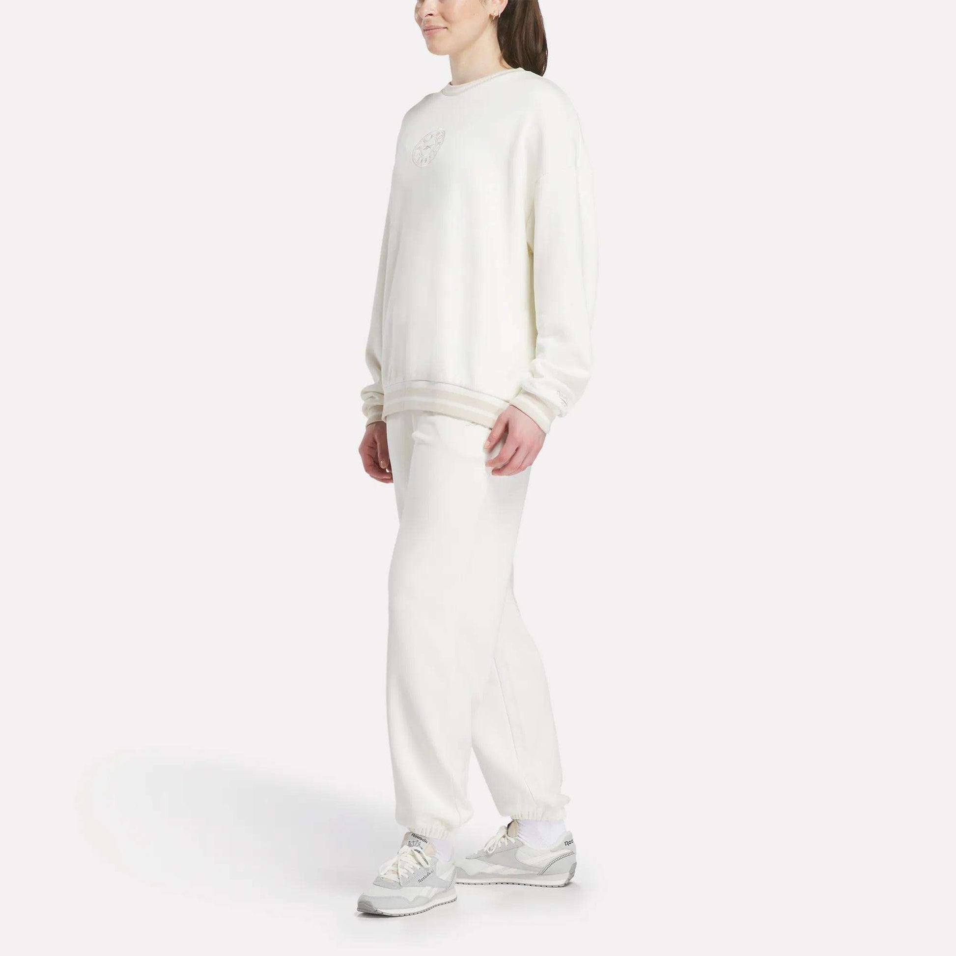 A person stands against a white background, wearing a loose-fitting Reebok Women's Lawn Club Crew Sweatshirt with matching white sweatpants and sneakers. Their hair is tied back, and their hands rest at their sides.