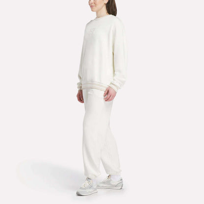 A person stands against a white background, wearing a loose-fitting Reebok Women's Lawn Club Crew Sweatshirt with matching white sweatpants and sneakers. Their hair is tied back, and their hands rest at their sides.