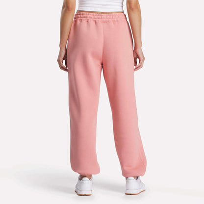 A person stands with their back to the camera against a plain white background, wearing pink Reebok Women's Wardrobe Essentials Sweat Pants, a white tank top, and white sneakers.
