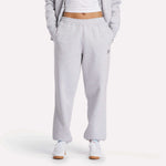 A person wearing Reebok Women's Wardrobe Essentials Sweat Pants with hands in pockets, paired with a white cropped top, white sneakers, and a matching gray jacket. The background is plain white.