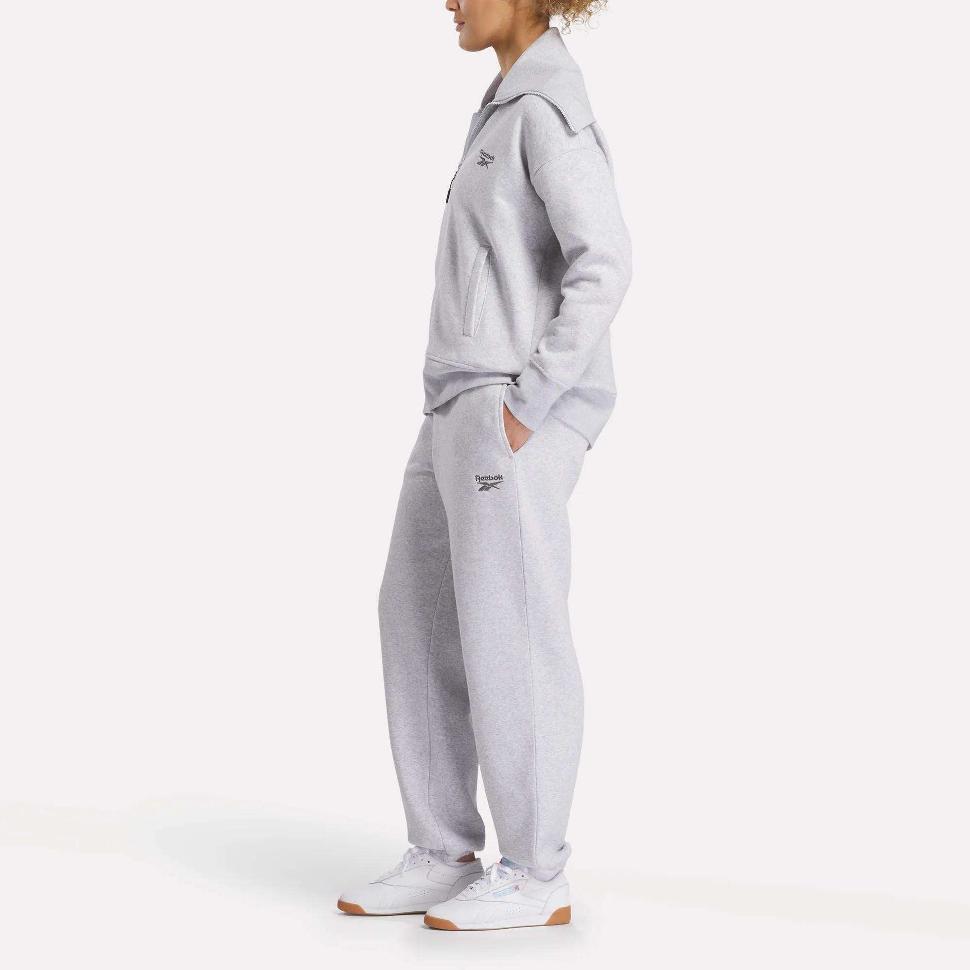 A person stands hands in pockets, wearing Reebok Women's Wardrobe Essentials Sweat Pants in light gray with a matching top and white sneakers. Small Reebok logos appear on the chest and leg as they face slightly to the side.