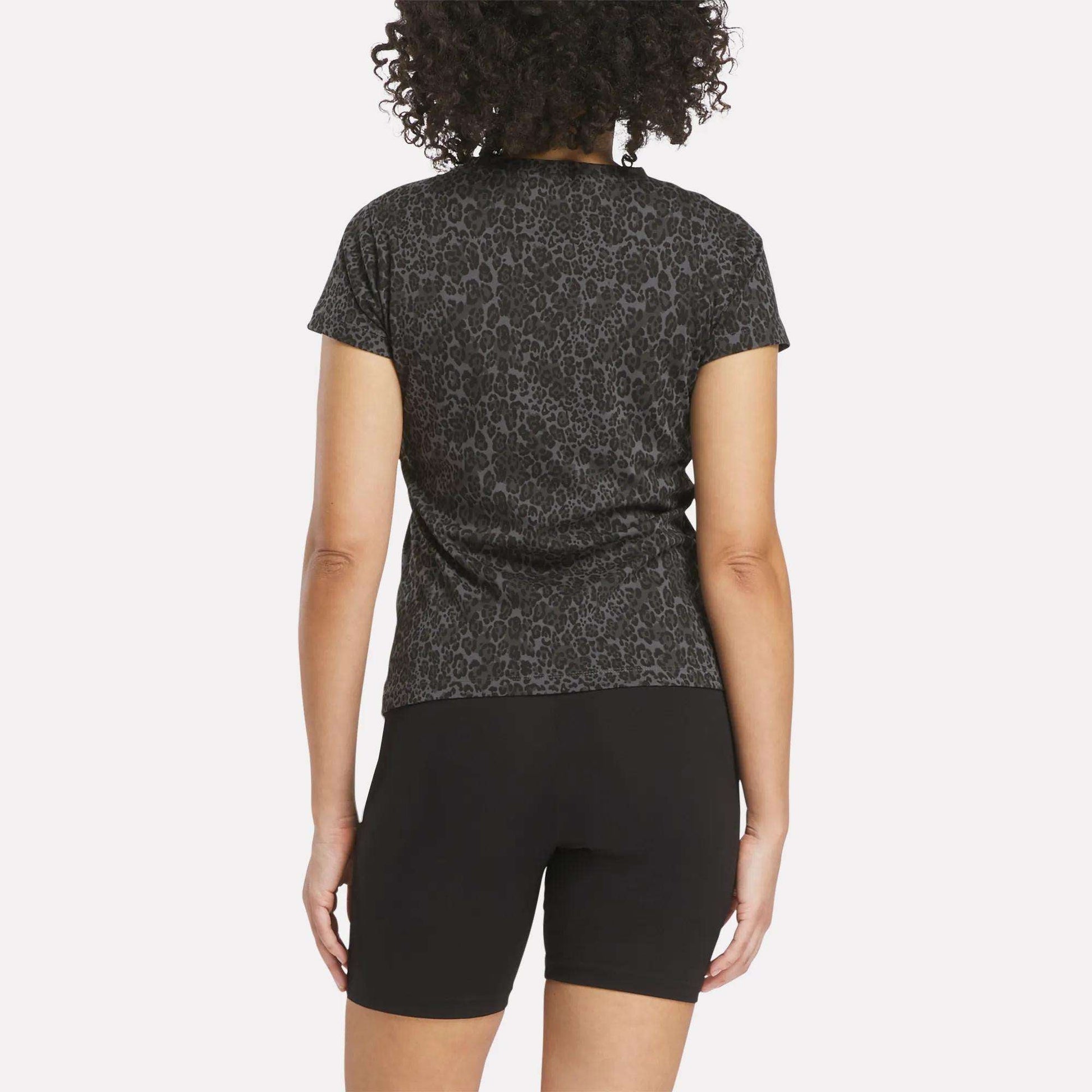 A person with curly hair is shown from the back wearing the Reebok Women's Leopard Print Baby T-Shirt and black shorts, posed against a plain white background.