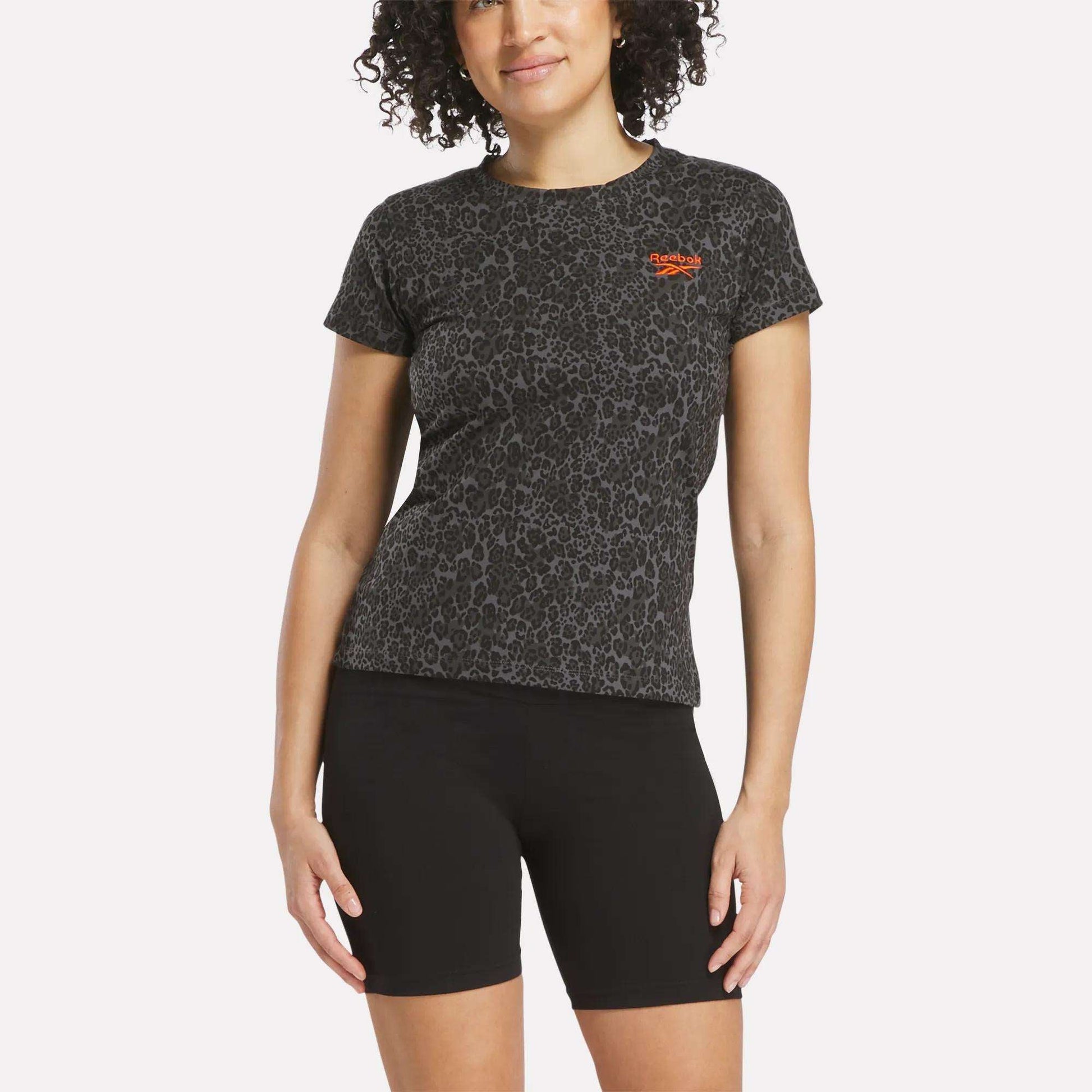 A woman with curly hair wears a black Reebok Women's Leopard Print Baby T-Shirt, featuring leopard print and an orange logo, paired with black bike shorts against a plain white background.