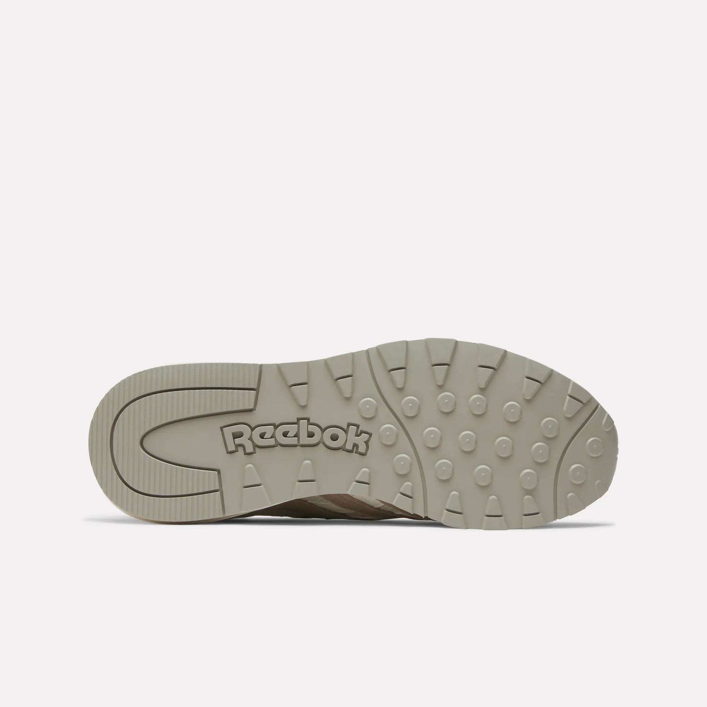 The image shows the beige rubber sole of the Reebok Women's Classic Nylon 89 Shoes, viewed from underneath. Circular grip patterns and an embossed Reebok logo near the heel highlight its retro design.