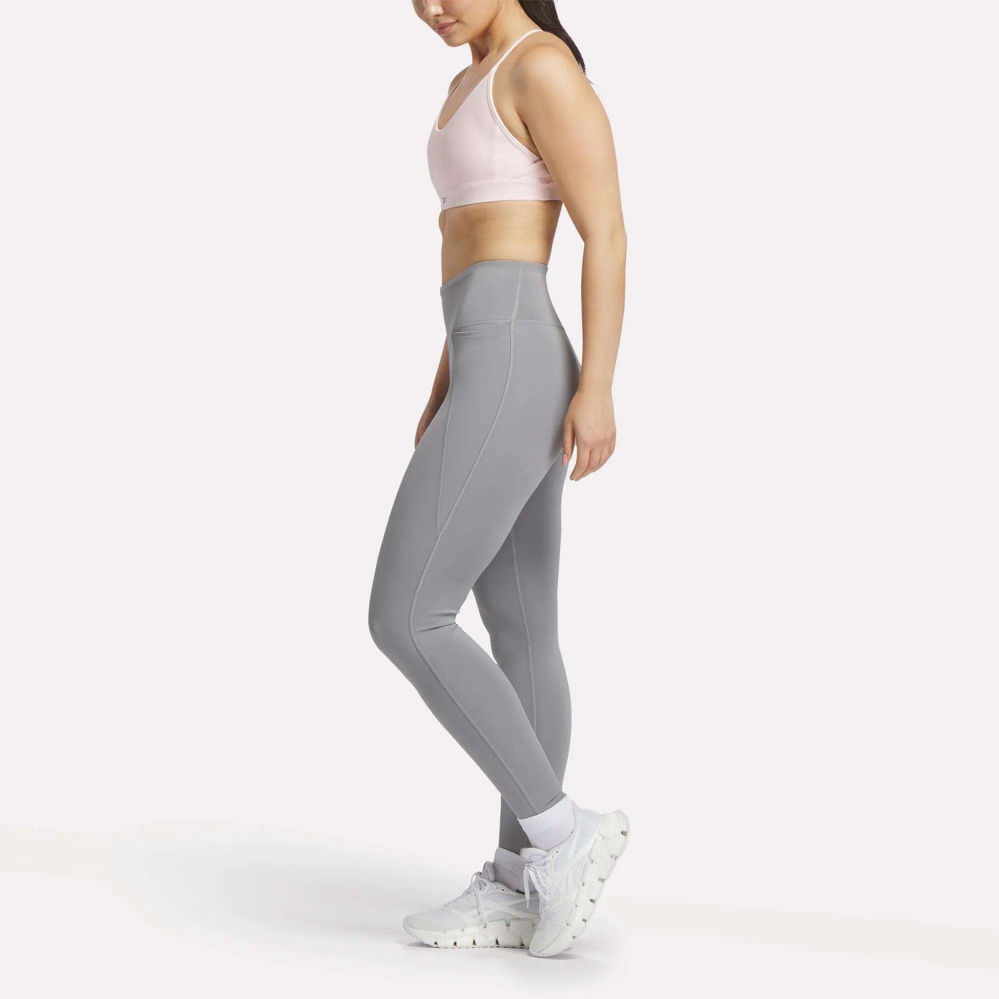 A woman stands sideways against a white background wearing Reebok Women's Lux-Lite High-Rise Tights, a light pink sports bra, white socks, and sneakers. The tights feature RBK-DRY performance fabric for comfort during training.