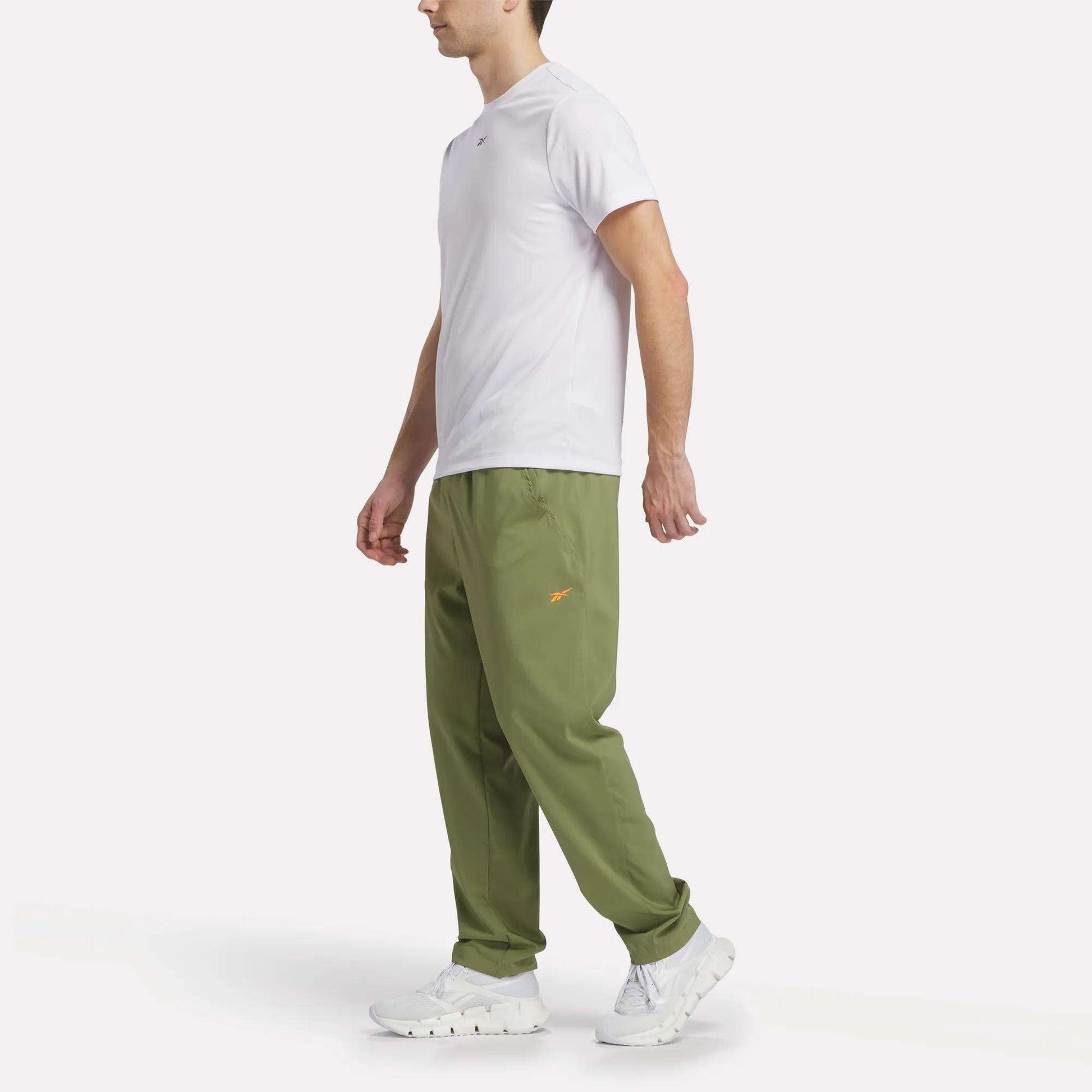 A man faces left against a plain white background, wearing a white t-shirt, white sneakers, and olive green Reebok Men's Train Woven Unlined Pants made from lightweight fabric with a small orange logo.