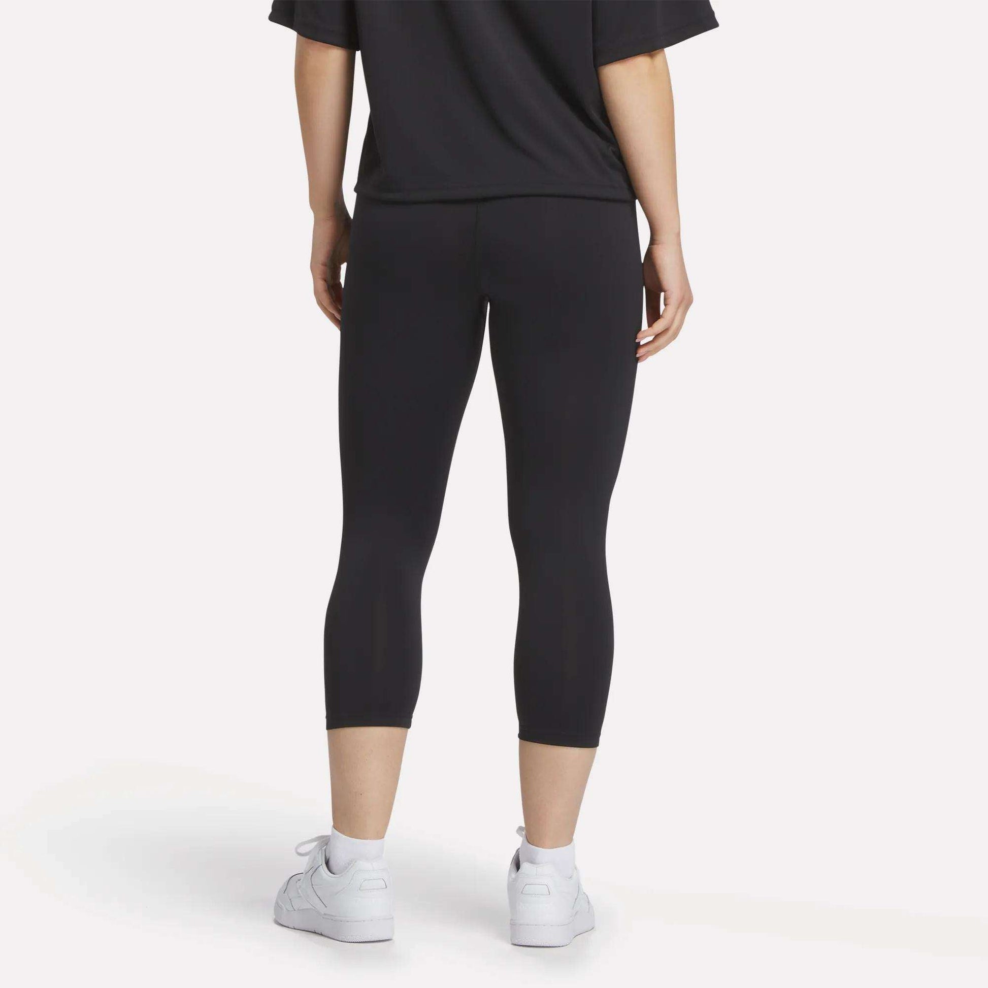 A person stands with their back to the camera, wearing a black t-shirt, white sneakers, and Reebok Women's ID Basketball 3/4 Compression Leggings against a plain light background.