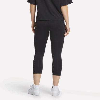 A person stands with their back to the camera, wearing a black t-shirt, white sneakers, and Reebok Women's ID Basketball 3/4 Compression Leggings against a plain light background.