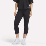 A figure from the shoulders down wears a loose black T-shirt, white sneakers, and Reebok Women's ID Basketball 3/4 Compression Leggings against a plain white background, highlighting the comfort and fit of these leggings.