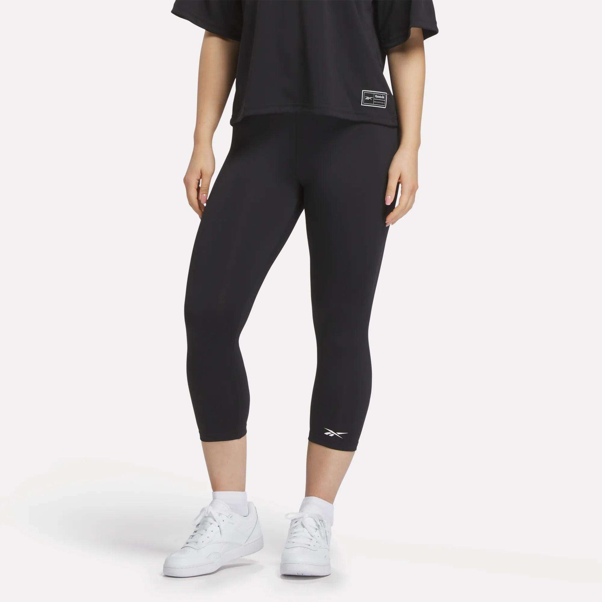 A figure from the shoulders down wears a loose black T-shirt, white sneakers, and Reebok Women's ID Basketball 3/4 Compression Leggings against a plain white background, highlighting the comfort and fit of these leggings.