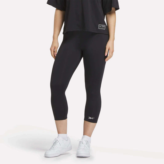 A figure from the shoulders down wears a loose black T-shirt, white sneakers, and Reebok Women's ID Basketball 3/4 Compression Leggings against a plain white background, highlighting the comfort and fit of these leggings.