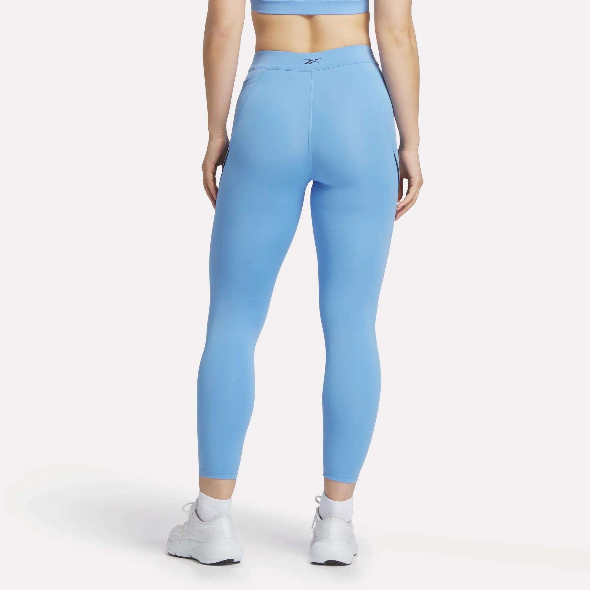 A woman stands facing away in Reebok Women's Lux High-Rise Colorblock Leggings and a light blue sports bra, paired with white athletic shoes. The plain white background highlights the comfort of the sweat-wicking fabric.