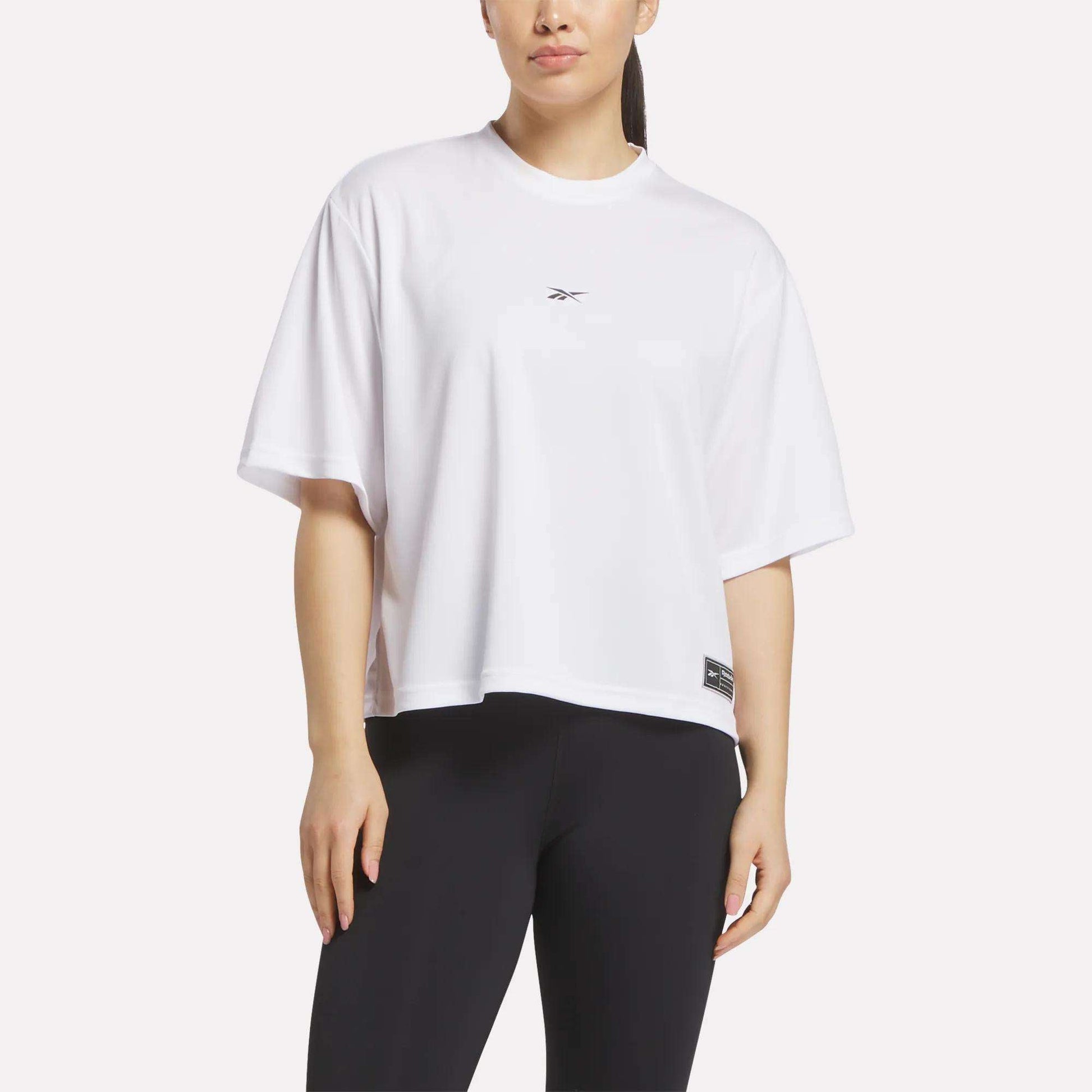 A woman stands against a light background in a loose-fitting Reebok Women's Basketball Mesh T-Shirt with a small chest logo and black leggings.