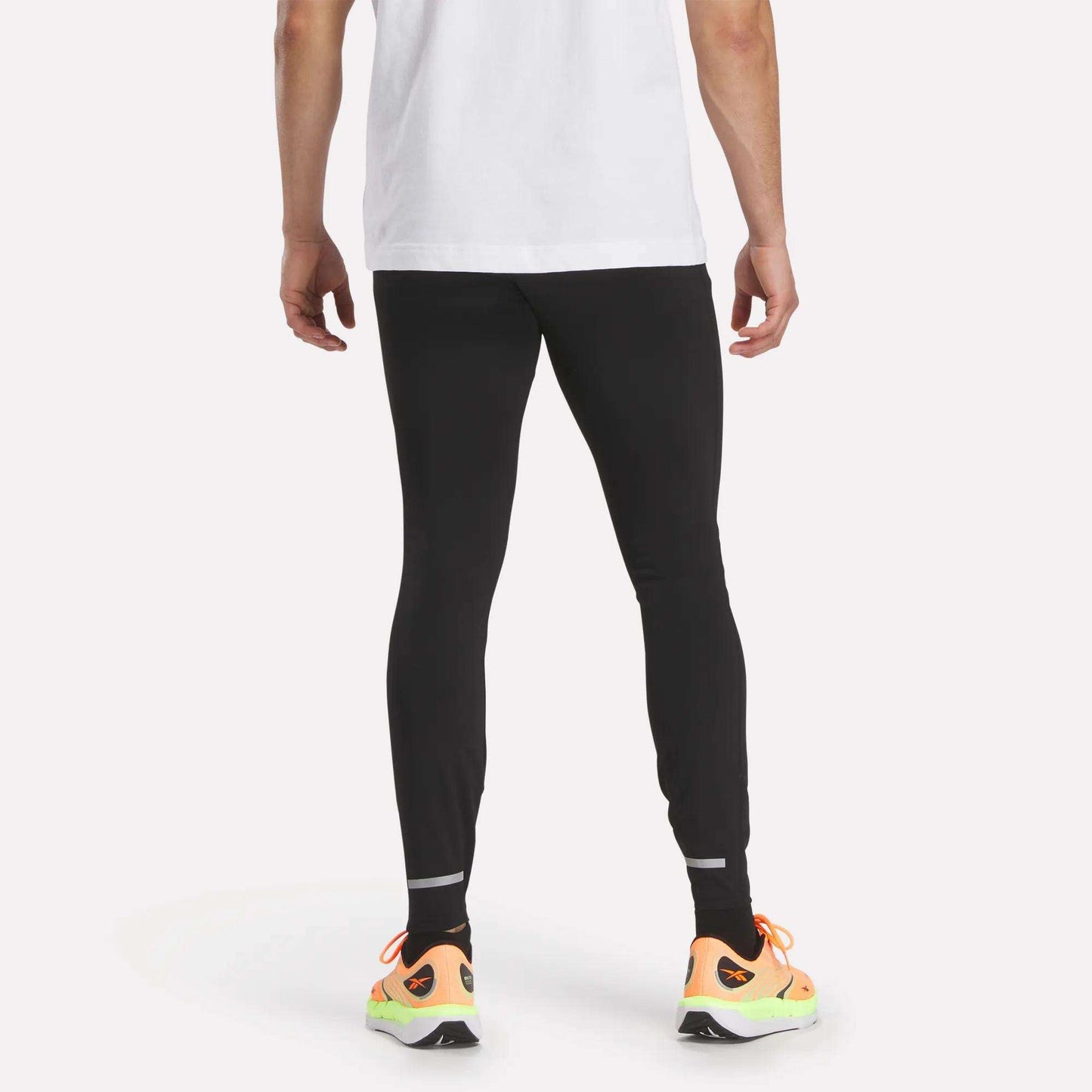 A person stands facing away in a white T-shirt, Reebok Men's Athlete Speed Running Tights, and bright orange sneakers with yellow soles against a plain white background.