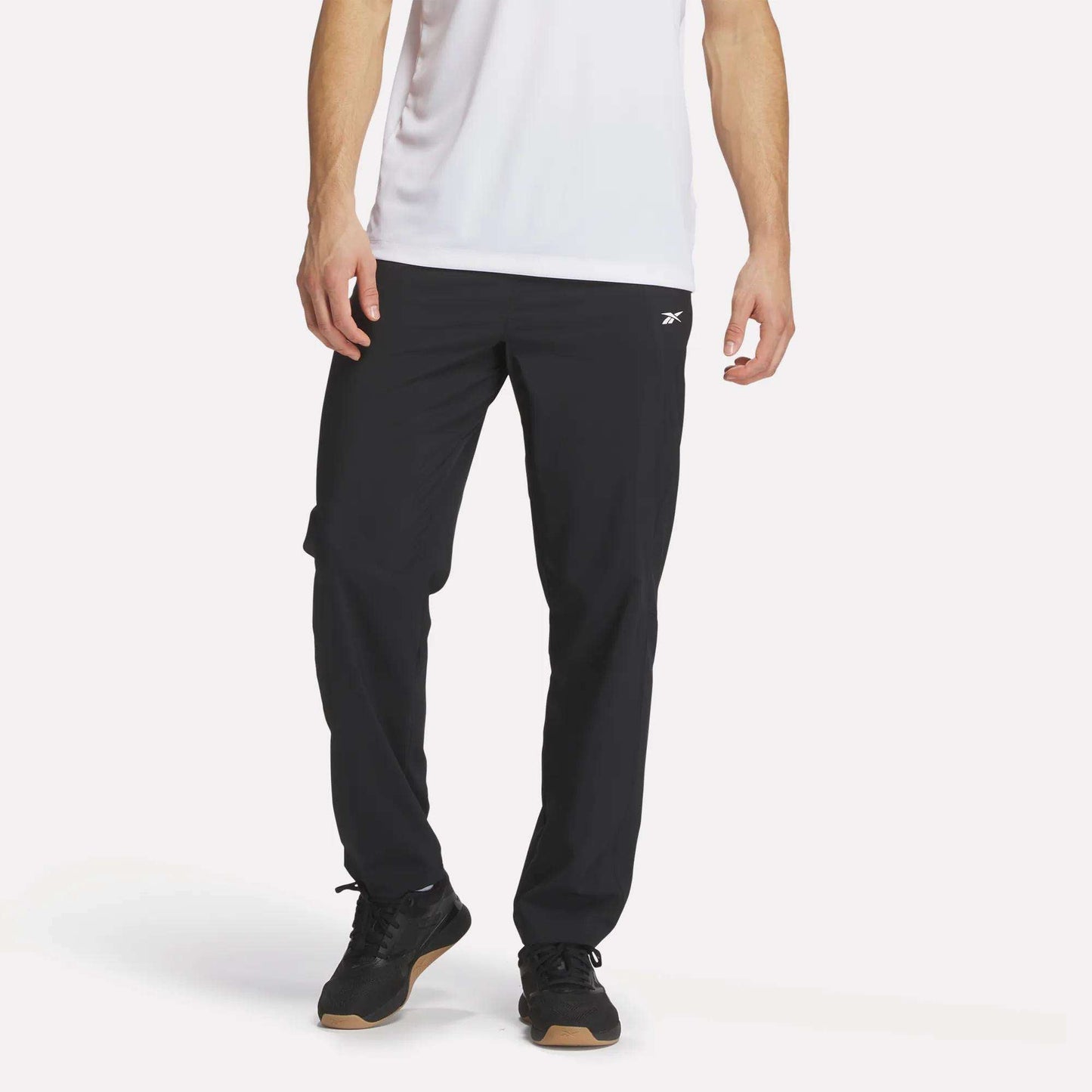 A person wears a white short-sleeve shirt and Reebok Men's Woven Pants, featuring a small white logo, with black sneakers, standing against a plain white background.