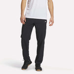 A person wears a white short-sleeve shirt and Reebok Men's Woven Pants, featuring a small white logo, with black sneakers, standing against a plain white background.