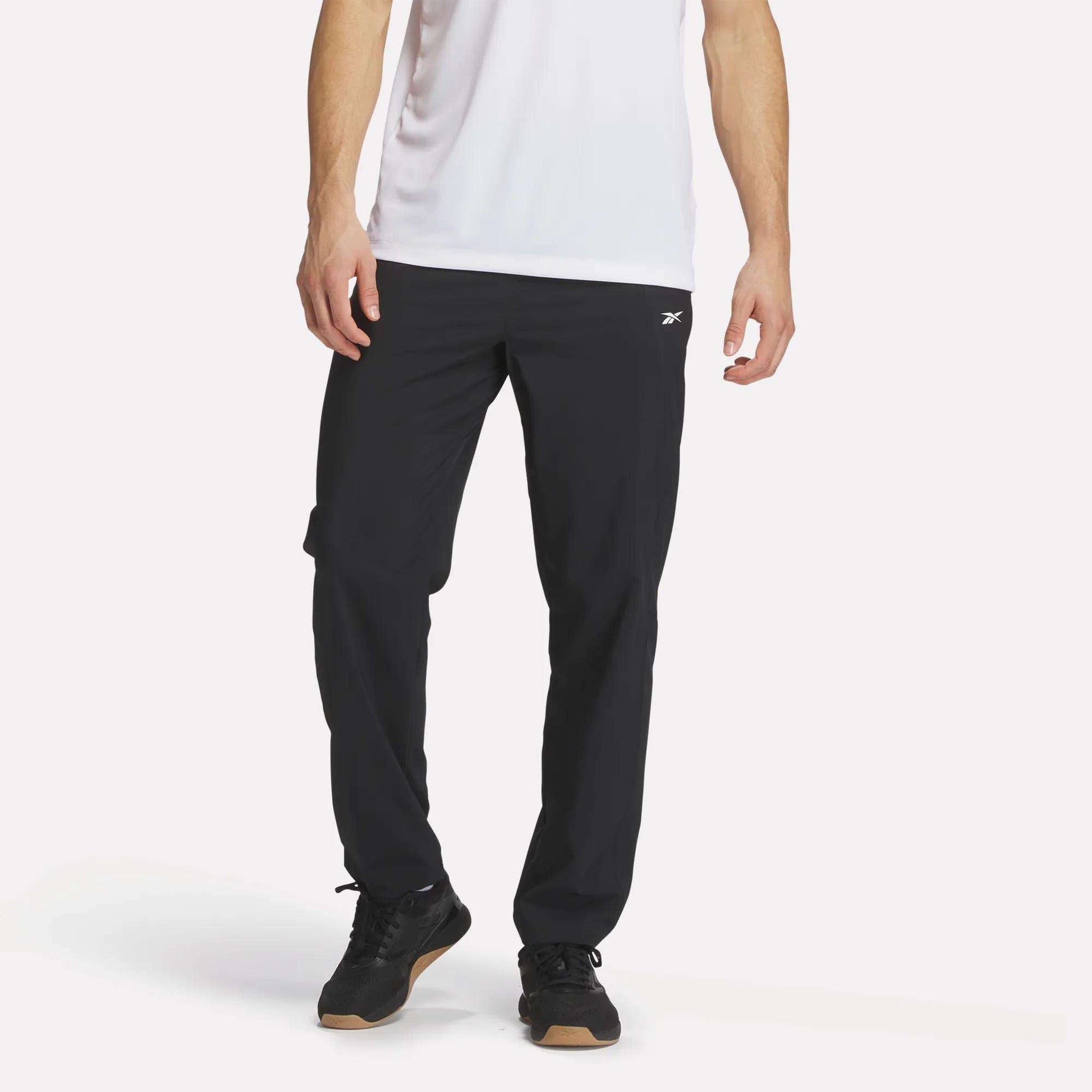 A person wears a white short-sleeve shirt and Reebok Men's Woven Pants, featuring a small white logo, with black sneakers, standing against a plain white background.