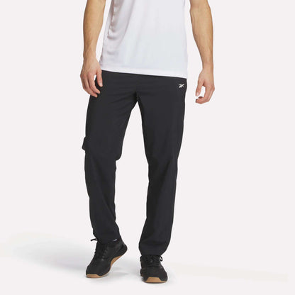 A person wears a white short-sleeve shirt and Reebok Men's Woven Pants, featuring a small white logo, with black sneakers, standing against a plain white background.