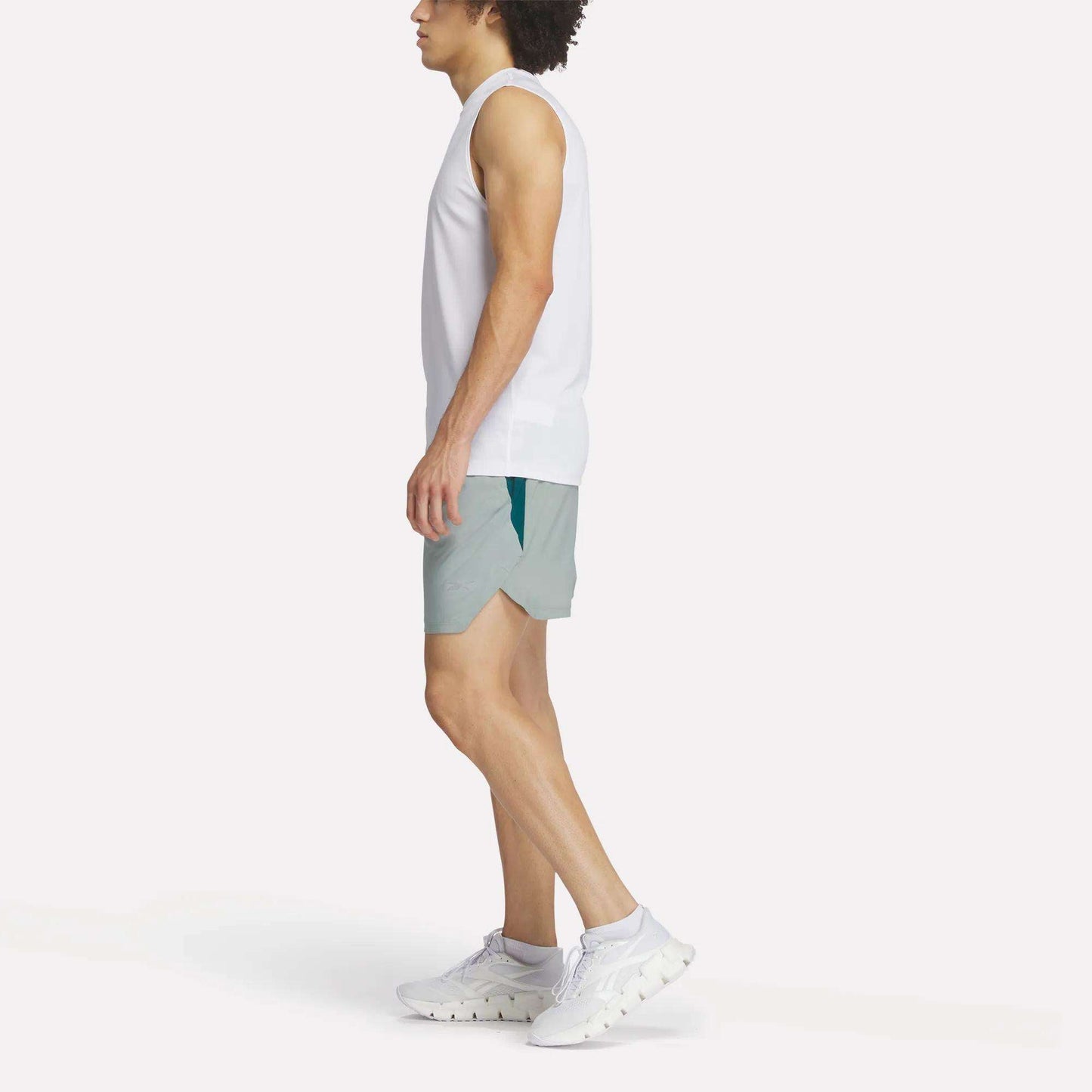 A person in a white sleeveless top, Reebok Men's Run Shorts with inner tights, and white sneakers stands sideways against a plain white background.