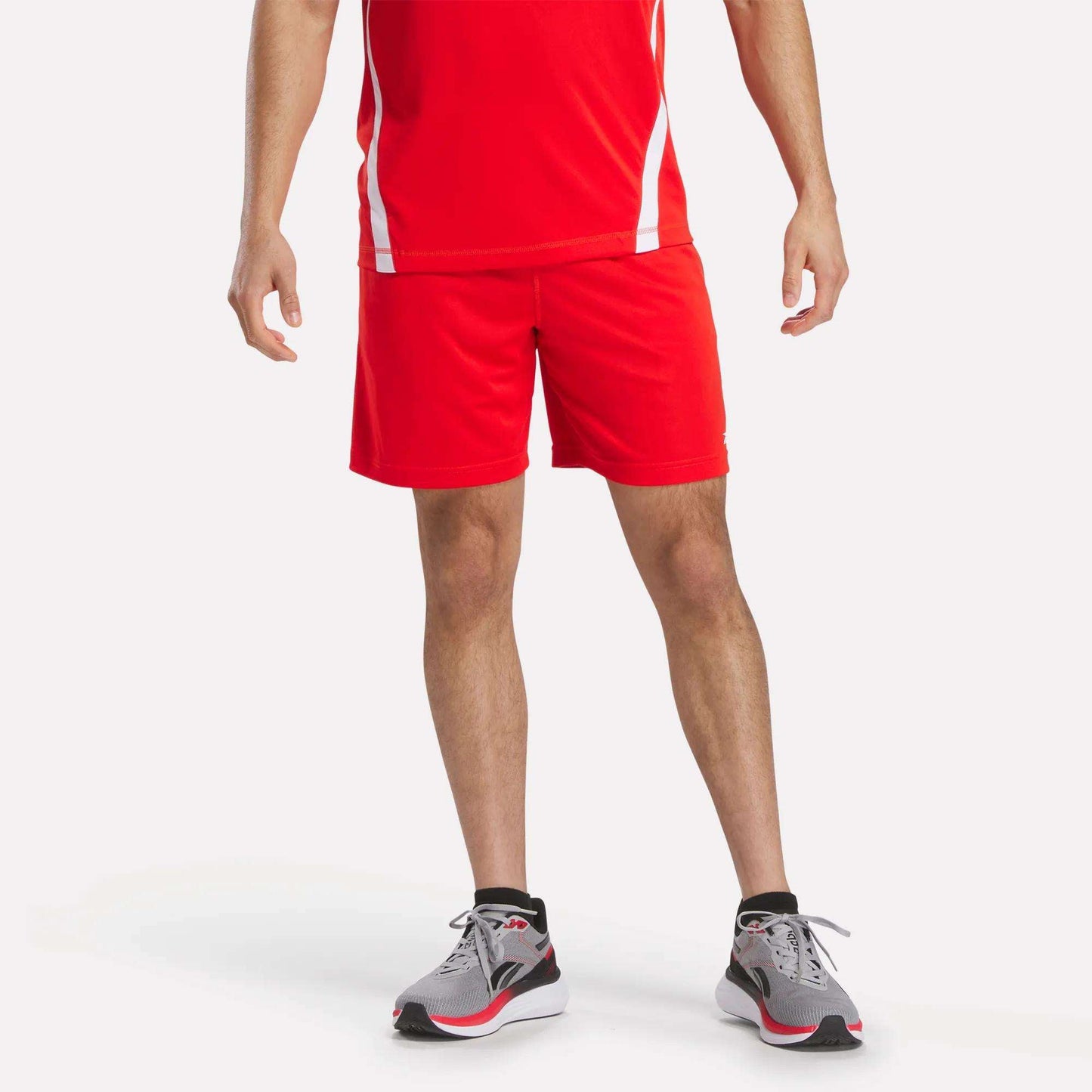 A person models Reebok Men's Train Blocked Knit Shorts, paired with a red athletic shirt and gray running shoes with red accents, against a plain white background. The head is cropped out—ideal for focusing on the workout wear.