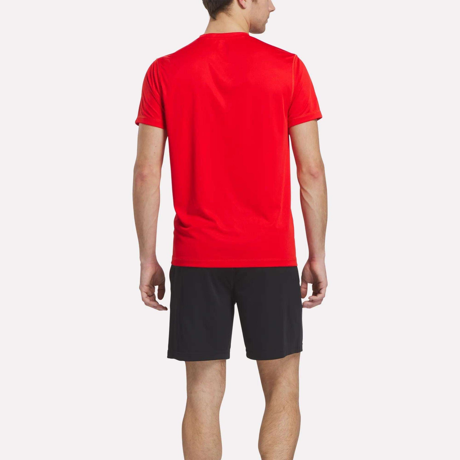 A man faces away from the camera, wearing a bright red Reebok Men's Train Blocked T-Shirt and black shorts against a plain, light background.